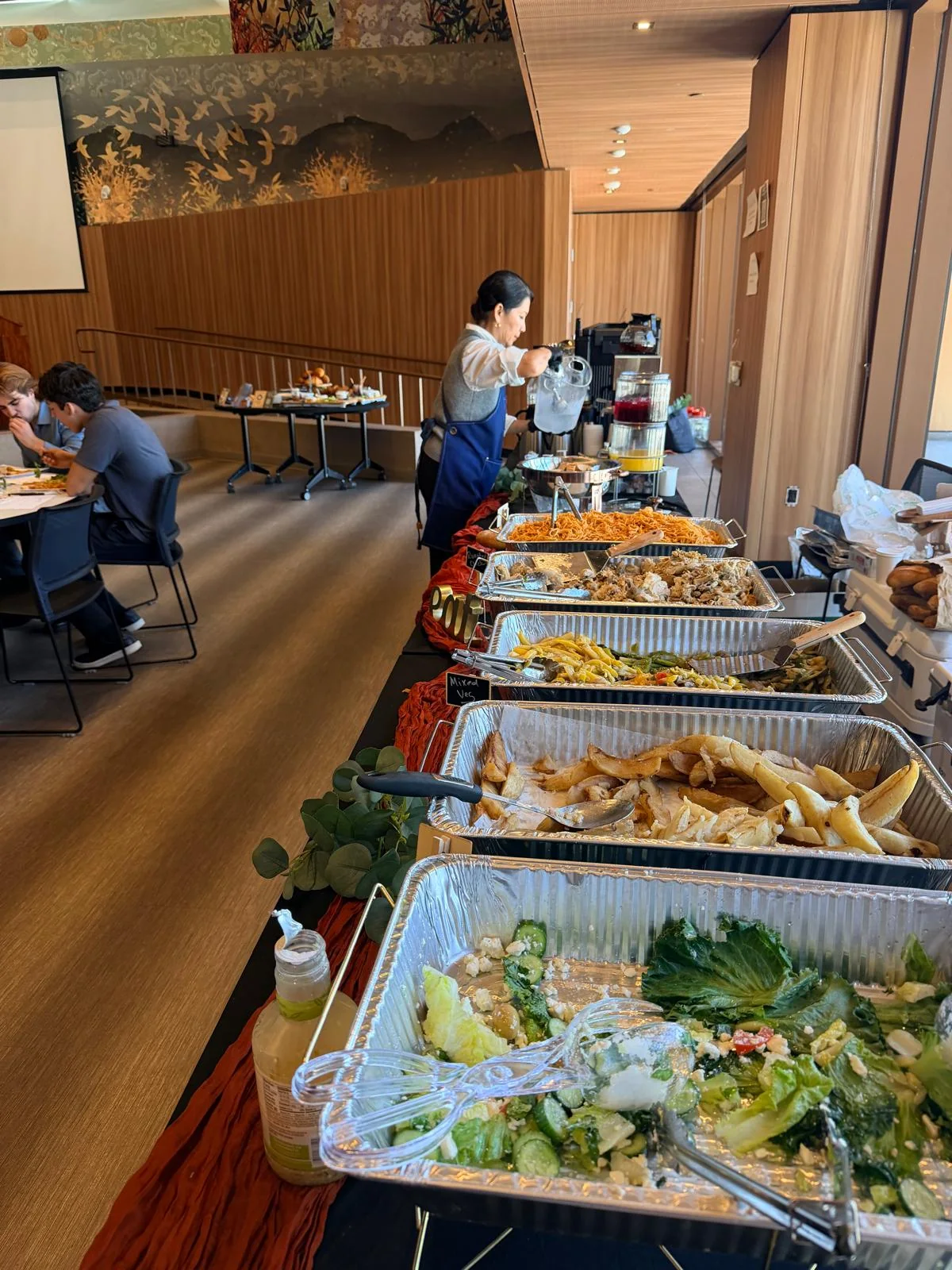 Midday hot lunch buffet line with chicken, pasta, grilled vegetables, and Greek salad